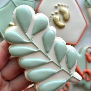 Fall-themed baby shower sugar cookies decorated with royal icing in sage green, burnt orange, ivory, and gold. Designs include pumpkins, “baby” lettering, baby onesies, bottles, bibs, pacifiers, leaves, and gold-accented baby footprints.
