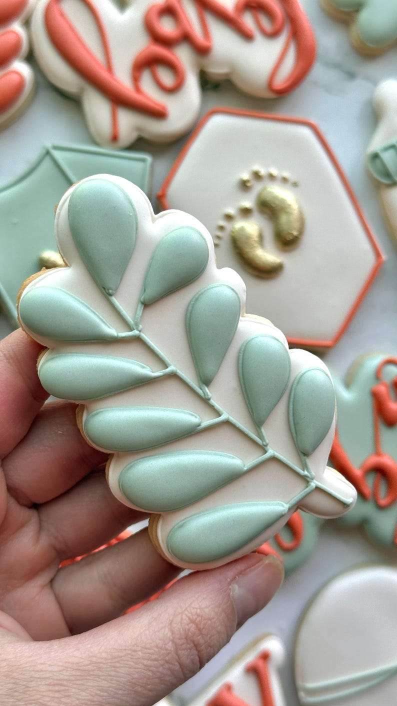 Fall-themed baby shower sugar cookies decorated with royal icing in sage green, burnt orange, ivory, and gold. Designs include pumpkins, “baby” lettering, baby onesies, bottles, bibs, pacifiers, leaves, and gold-accented baby footprints.