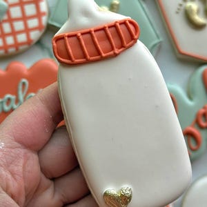 Fall-themed baby shower sugar cookies decorated with royal icing in sage green, burnt orange, ivory, and gold. Designs include pumpkins, “baby” lettering, baby onesies, bottles, bibs, pacifiers, leaves, and gold-accented baby footprints.