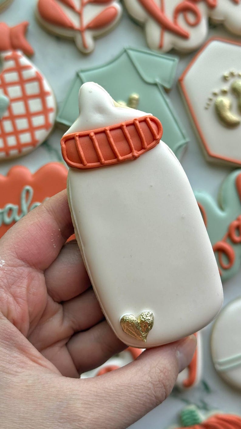 Fall-themed baby shower sugar cookies decorated with royal icing in sage green, burnt orange, ivory, and gold. Designs include pumpkins, “baby” lettering, baby onesies, bottles, bibs, pacifiers, leaves, and gold-accented baby footprints.