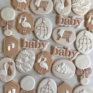 May include: A collection of 19 decorated sugar cookies with a baby shower theme. The cookies are decorated with brown and white icing and feature images of storks, baby mobiles, clouds, and the word "baby".