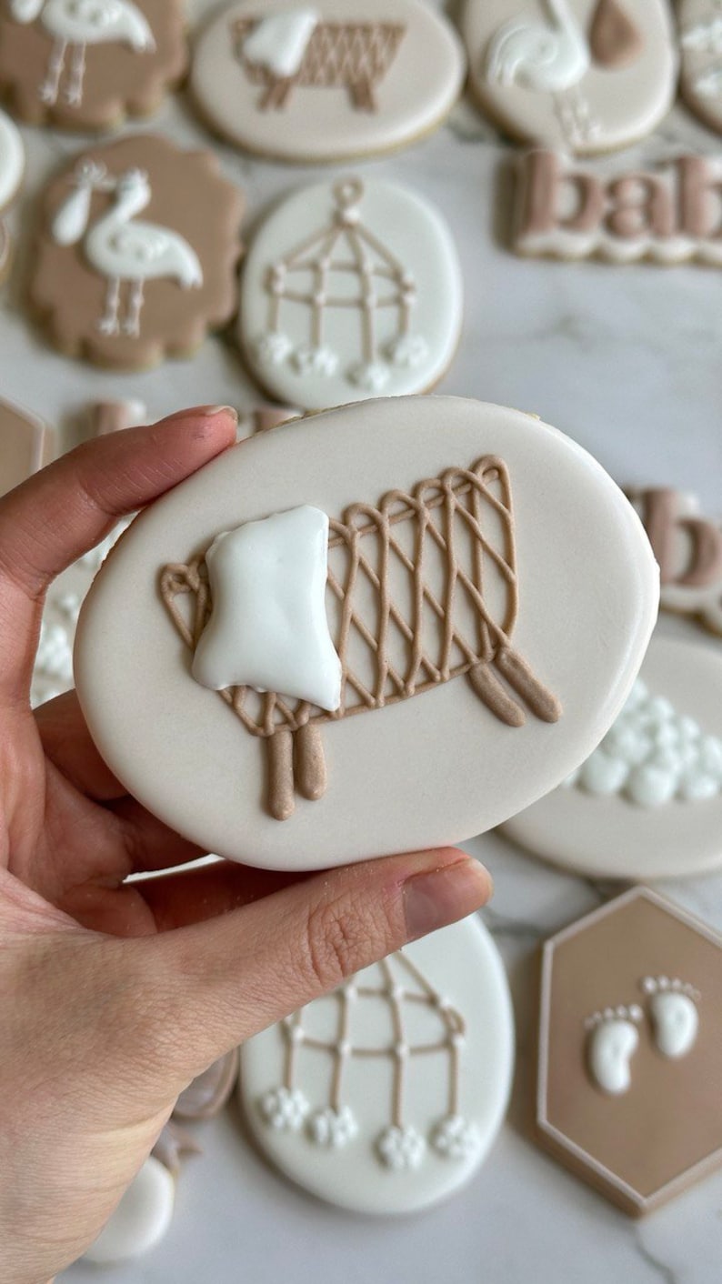 May include: A collection of decorated sugar cookies with various baby-themed designs, including a crib, a mobile, a stork, and footprints. The cookies are decorated with white and brown icing.