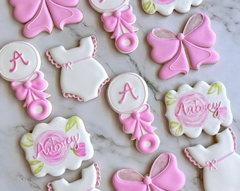 May include: A set of twelve pink and white baby shower cookies. The cookies are decorated with bows, rattles, onesies, and the name "Aubrey".