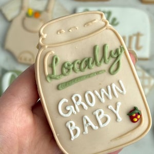 Farmers market-themed baby shower sugar cookies featuring hand-decorated designs like “little sprout,” baby onesies with vegetables, carrots, corn, strawberries, tomatoes, baby bottles, mason jars, leafy greens, and baby mobiles. Adorned with soft green, beige, orange, and red royal icing, these custom cookies are perfect for a “locally grown” or garden baby shower theme, gender-neutral celebrations, and personalized baby gifts.