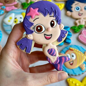 Może przedstawiać: Zestaw ozdobnych ciasteczek cukrowych z postaciami z dziecięcego programu telewizyjnego Bubble Guppies. Ciasteczka są ozdobione kolorową polewą i przedstawiają postacie takie jak Molly, Gil i Deema.