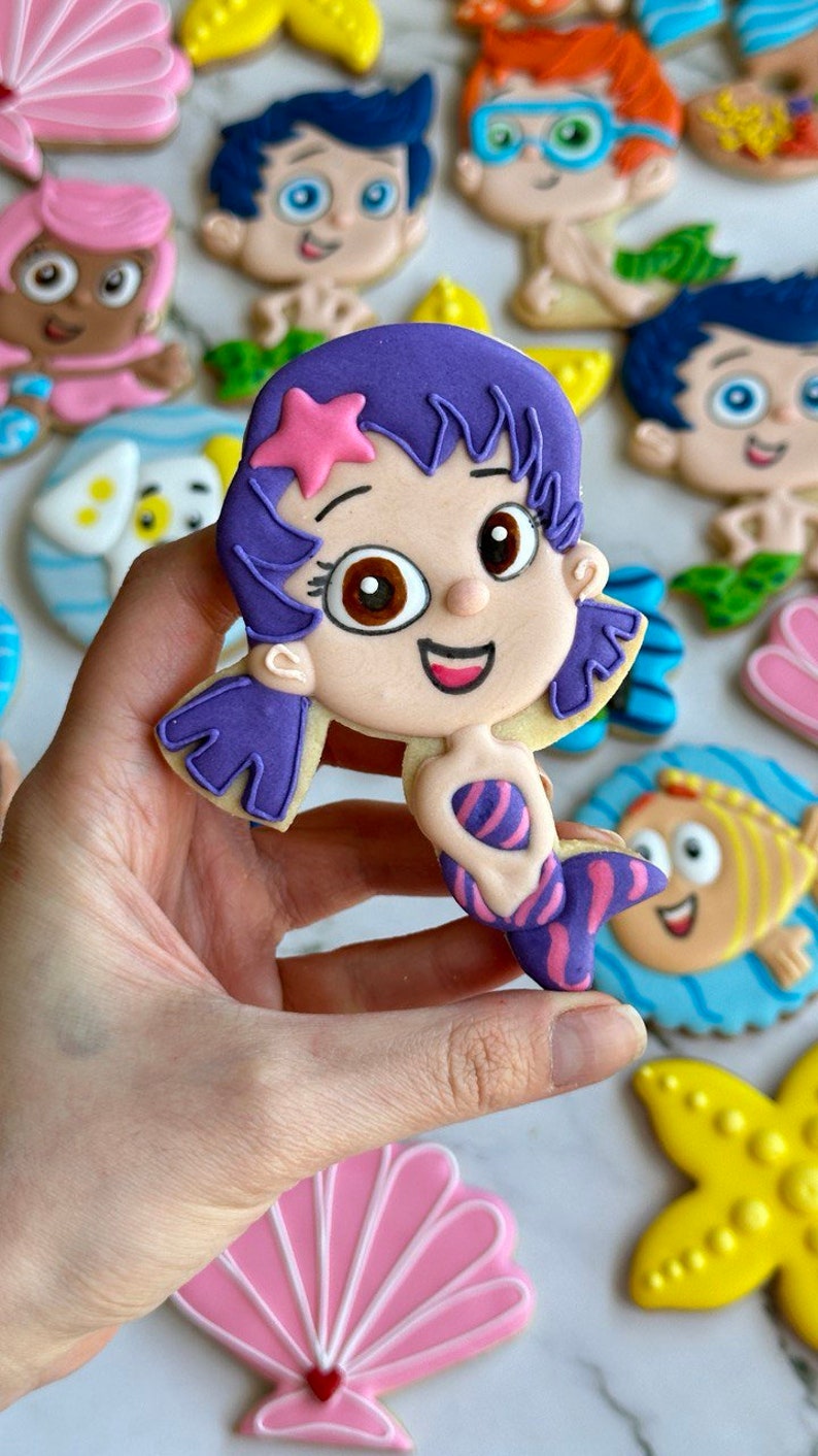 Może przedstawiać: Zestaw ozdobnych ciasteczek cukrowych z postaciami z dziecięcego programu telewizyjnego Bubble Guppies. Ciasteczka są ozdobione kolorową polewą i przedstawiają postacie takie jak Molly, Gil i Deema.