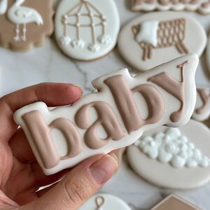 May include: A collection of decorated sugar cookies with various baby themes, including a stork, a crib mobile, a sheep, and the word "baby" in brown icing.