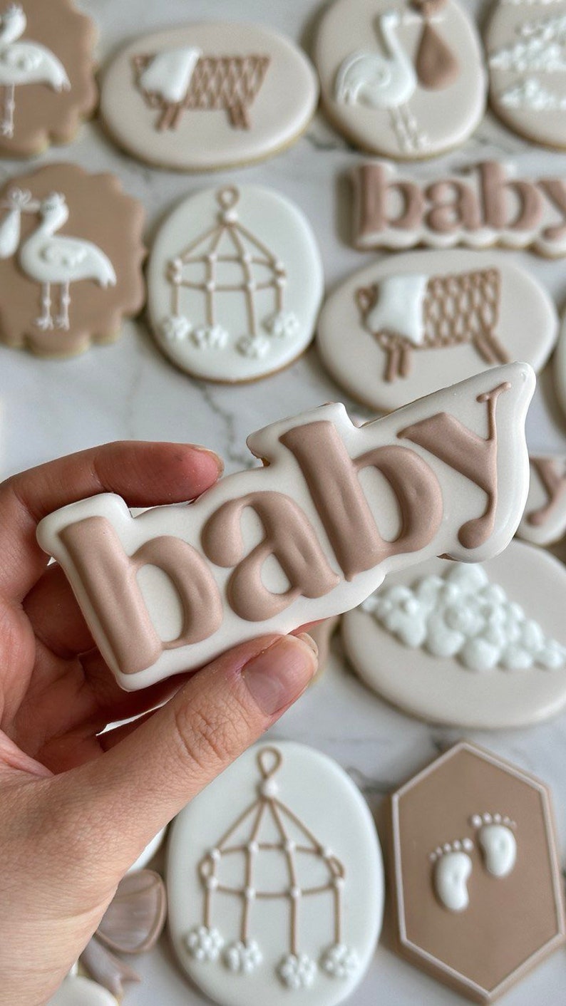 May include: A collection of decorated sugar cookies with various baby themes, including a stork, a crib mobile, a sheep, and the word "baby" in brown icing.