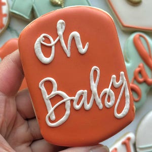 Fall-themed baby shower sugar cookies decorated with royal icing in sage green, burnt orange, ivory, and gold. Designs include pumpkins, “baby” lettering, baby onesies, bottles, bibs, pacifiers, leaves, and gold-accented baby footprints.