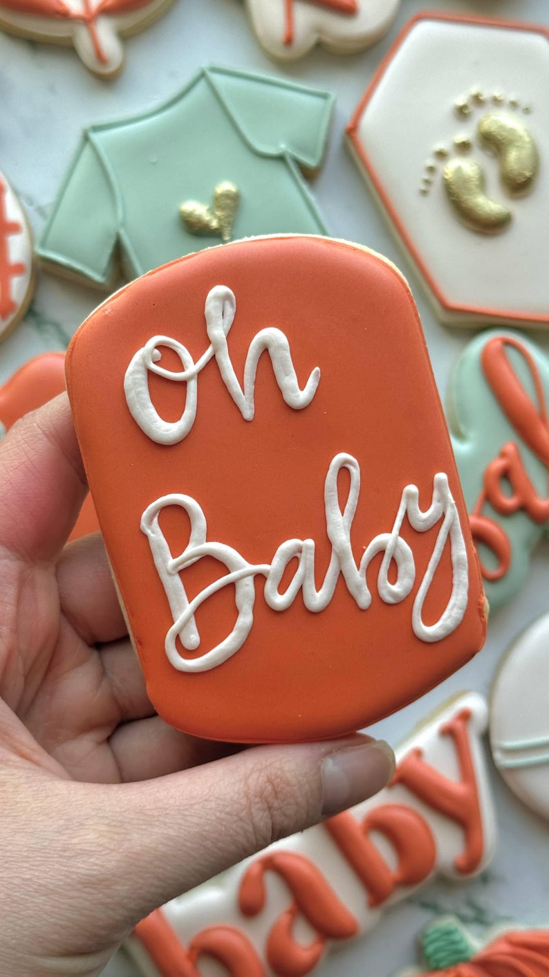 Fall-themed baby shower sugar cookies decorated with royal icing in sage green, burnt orange, ivory, and gold. Designs include pumpkins, “baby” lettering, baby onesies, bottles, bibs, pacifiers, leaves, and gold-accented baby footprints.