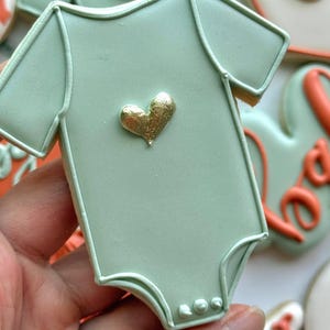 Fall-themed baby shower sugar cookies decorated with royal icing in sage green, burnt orange, ivory, and gold. Designs include pumpkins, “baby” lettering, baby onesies, bottles, bibs, pacifiers, leaves, and gold-accented baby footprints.