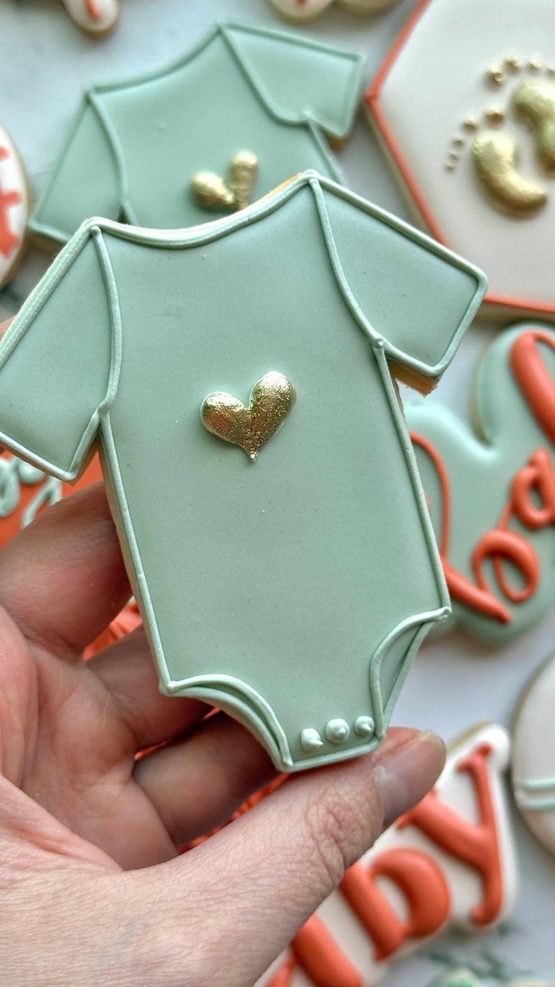 Fall-themed baby shower sugar cookies decorated with royal icing in sage green, burnt orange, ivory, and gold. Designs include pumpkins, “baby” lettering, baby onesies, bottles, bibs, pacifiers, leaves, and gold-accented baby footprints.