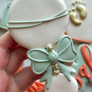Fall-themed baby shower sugar cookies decorated with royal icing in sage green, burnt orange, ivory, and gold. Designs include pumpkins, “baby” lettering, baby onesies, bottles, bibs, pacifiers, leaves, and gold-accented baby footprints.
