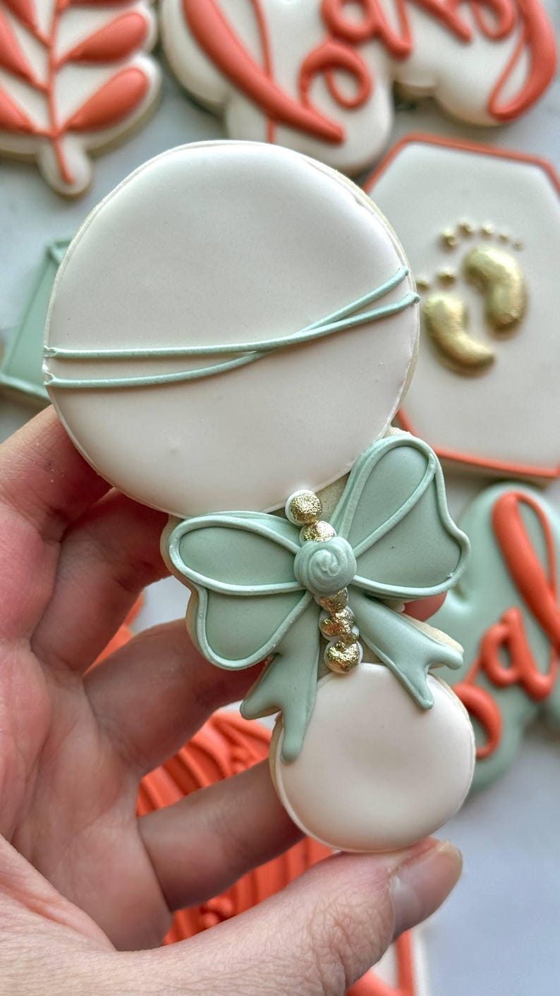 Fall-themed baby shower sugar cookies decorated with royal icing in sage green, burnt orange, ivory, and gold. Designs include pumpkins, “baby” lettering, baby onesies, bottles, bibs, pacifiers, leaves, and gold-accented baby footprints.
