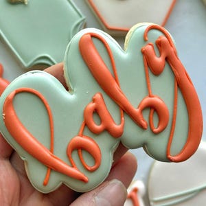 Fall-themed baby shower sugar cookies decorated with royal icing in sage green, burnt orange, ivory, and gold. Designs include pumpkins, “baby” lettering, baby onesies, bottles, bibs, pacifiers, leaves, and gold-accented baby footprints.