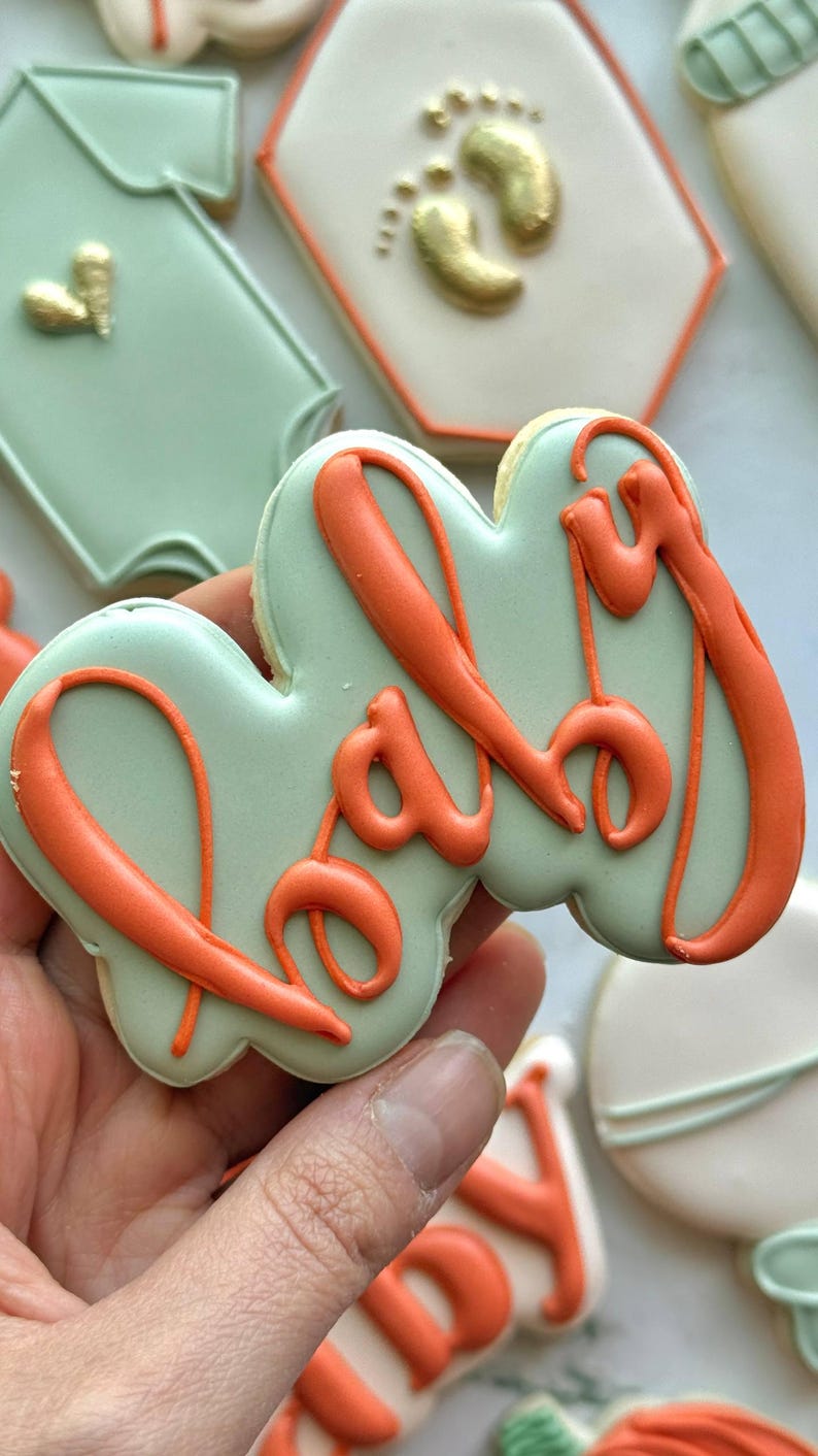 Fall-themed baby shower sugar cookies decorated with royal icing in sage green, burnt orange, ivory, and gold. Designs include pumpkins, “baby” lettering, baby onesies, bottles, bibs, pacifiers, leaves, and gold-accented baby footprints.