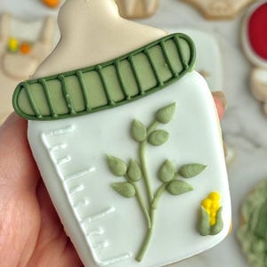 Farmers market-themed baby shower sugar cookies featuring hand-decorated designs like “little sprout,” baby onesies with vegetables, carrots, corn, strawberries, tomatoes, baby bottles, mason jars, leafy greens, and baby mobiles. Adorned with soft green, beige, orange, and red royal icing, these custom cookies are perfect for a “locally grown” or garden baby shower theme, gender-neutral celebrations, and personalized baby gifts.