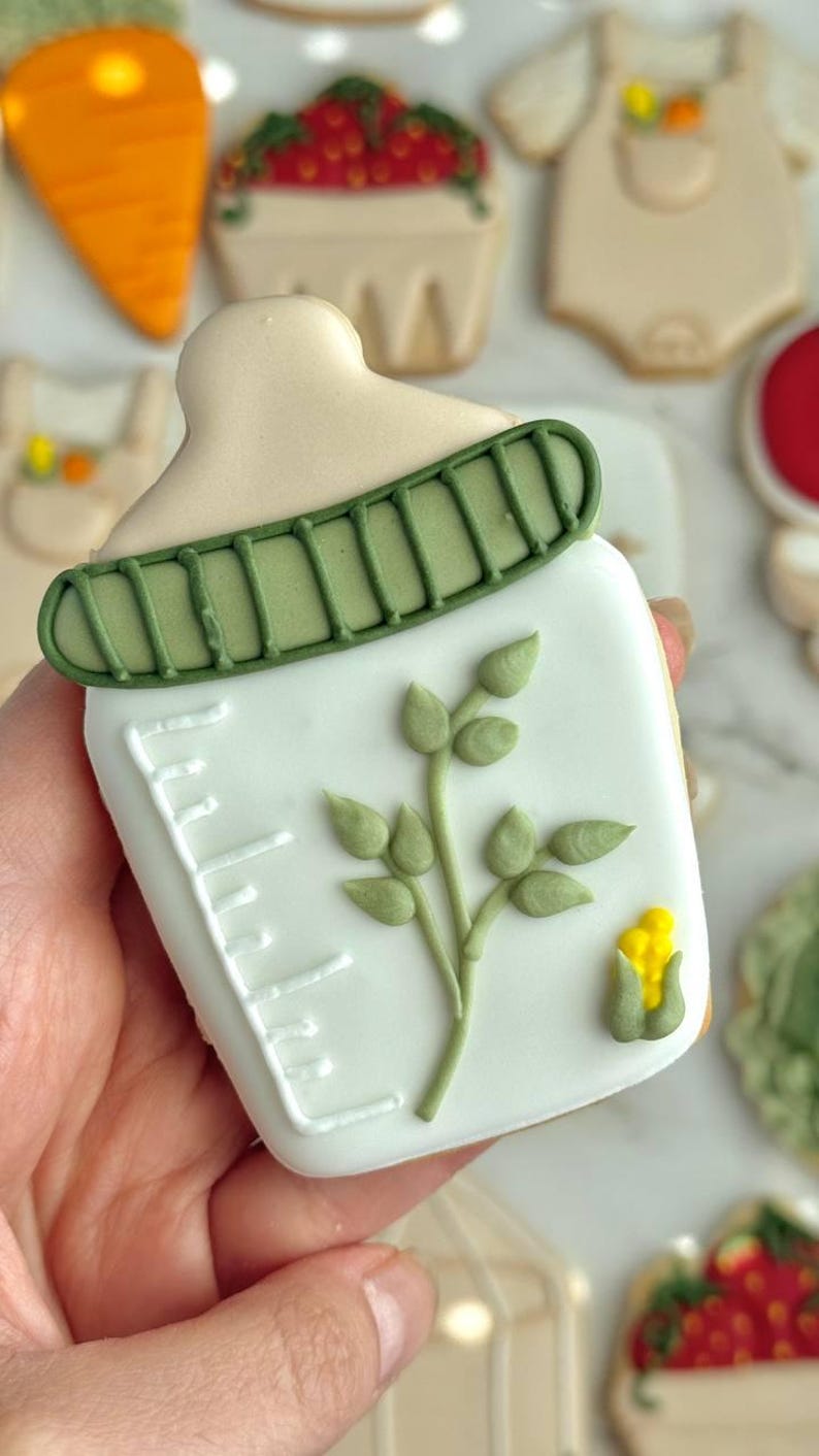 Farmers market-themed baby shower sugar cookies featuring hand-decorated designs like “little sprout,” baby onesies with vegetables, carrots, corn, strawberries, tomatoes, baby bottles, mason jars, leafy greens, and baby mobiles. Adorned with soft green, beige, orange, and red royal icing, these custom cookies are perfect for a “locally grown” or garden baby shower theme, gender-neutral celebrations, and personalized baby gifts.