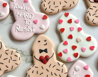 May include: A collection of penis-shaped cookies, decorated with white, pink, and tan icing. Some cookies have heart-shaped sprinkles, while others feature bow ties and heart details. One cookie reads "You're Hard To Resist."