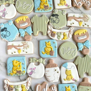 May include: A collection of Winnie the Pooh themed sugar cookies decorated with various designs, including Winnie the Pooh holding a honey pot, a baby bottle, a bib, and the words "Oh Boy" and "Baby".