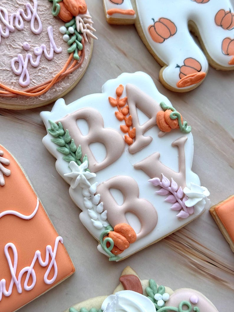 Assorted boho pumpkin baby shower sugar cookies decorated with pumpkins, baby onesie, baby bottle, rainbow, baby feet, “Little Pumpkin,” “Oh Baby,” “Baby Girl,” and “BABY” plaque in soft blush, sage, ivory, and pumpkin orange.