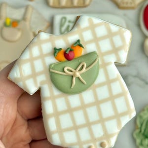 Farmers market-themed baby shower sugar cookies featuring hand-decorated designs like “little sprout,” baby onesies with vegetables, carrots, corn, strawberries, tomatoes, baby bottles, mason jars, leafy greens, and baby mobiles. Adorned with soft green, beige, orange, and red royal icing, these custom cookies are perfect for a “locally grown” or garden baby shower theme, gender-neutral celebrations, and personalized baby gifts.