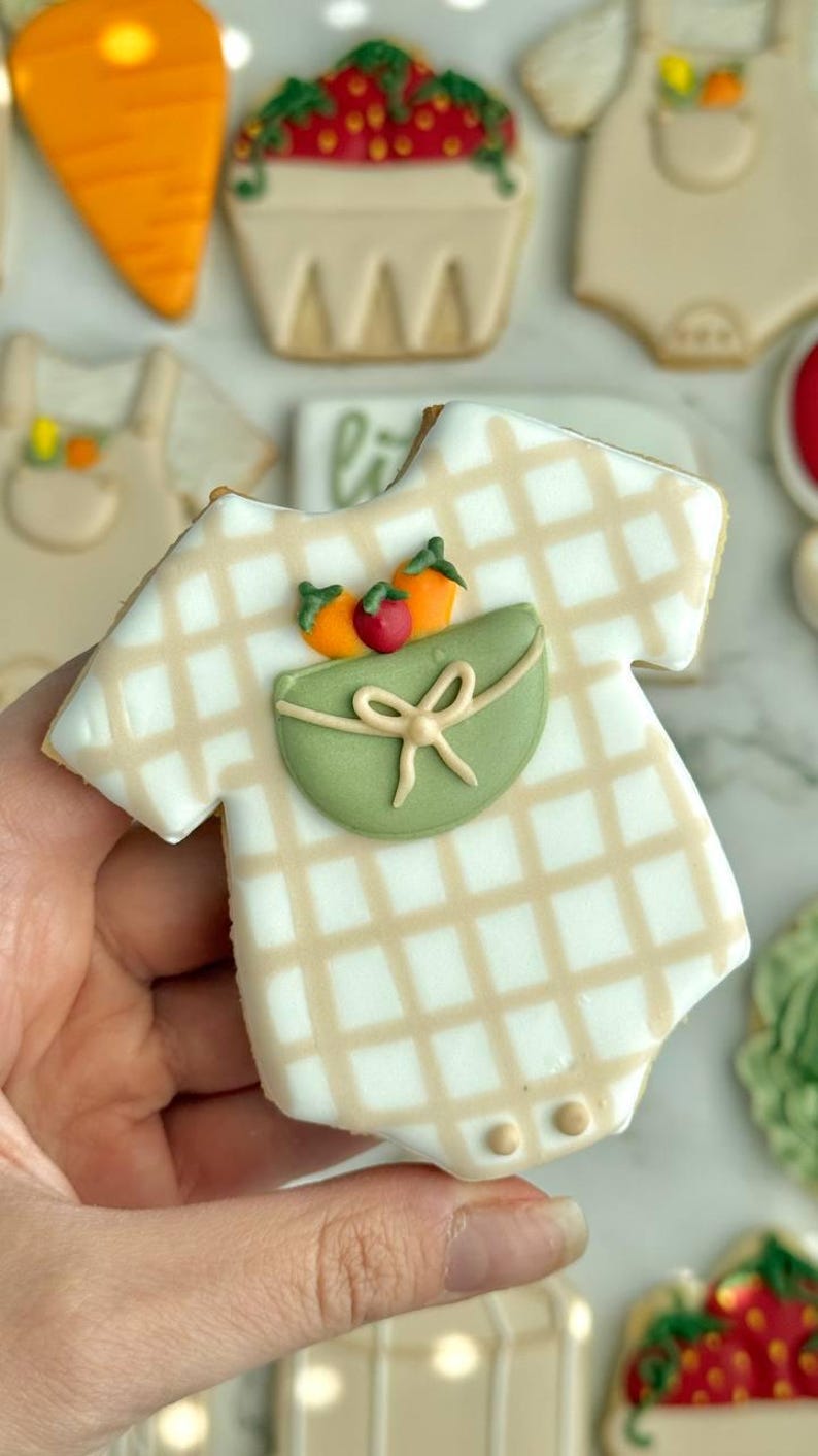 Farmers market-themed baby shower sugar cookies featuring hand-decorated designs like “little sprout,” baby onesies with vegetables, carrots, corn, strawberries, tomatoes, baby bottles, mason jars, leafy greens, and baby mobiles. Adorned with soft green, beige, orange, and red royal icing, these custom cookies are perfect for a “locally grown” or garden baby shower theme, gender-neutral celebrations, and personalized baby gifts.
