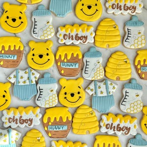 May include: A collection of Winnie the Pooh-themed biscuits. The biscuits feature smiling bear faces, honey pots with "HUNNY" text, baby bottles, beehives, and baby onesies with bee designs. The phrase "oh boy" is also featured on some biscuits.