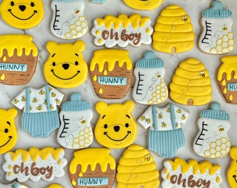 May include: Assortment of Winnie the Pooh-themed sugar cookies. The cookies include smiling bear faces, honey pots with "HUNNY" text, baby bottles, beehives, and baby onesies with bee designs. The phrase "oh boy" is also featured on some cookies.