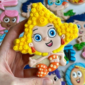 Może przedstawiać: Zestaw kolorowych, ozdobnych ciasteczek cukrowych z postaciami z dziecięcego programu telewizyjnego Bubble Guppies. Ciasteczka są ozdobione ż&oacute;łtą, niebieską, pomarańczową, r&oacute;żową i fioletową polewą. Ciasteczka mają kształt ryb, muszli i gwiazd.