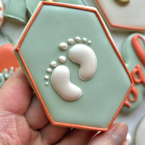 Fall-themed baby shower sugar cookies decorated with royal icing in sage green, burnt orange, ivory, and gold. Designs include pumpkins, “baby” lettering, baby onesies, bottles, bibs, pacifiers, leaves, and gold-accented baby footprints.