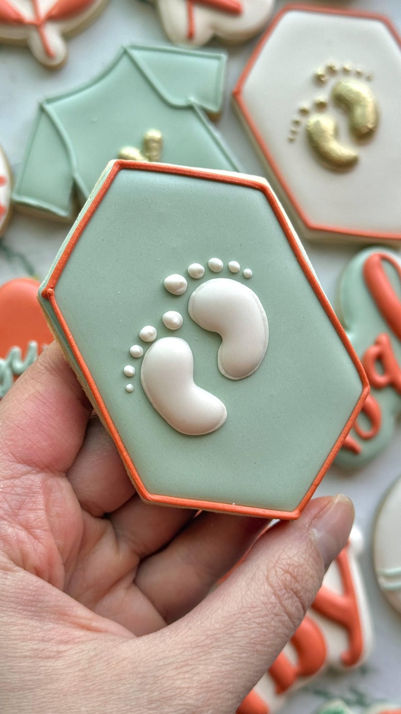 Fall-themed baby shower sugar cookies decorated with royal icing in sage green, burnt orange, ivory, and gold. Designs include pumpkins, “baby” lettering, baby onesies, bottles, bibs, pacifiers, leaves, and gold-accented baby footprints.