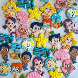 Może przedstawiać: Kolekcja kolorowych, ozdobnych ciasteczek cukrowych z postaciami z dziecięcego programu telewizyjnego Bubble Guppies. Ciasteczka mają kształt ryb, muszli, rozgwiazd i innych obiekt&oacute;w o tematyce oceanicznej. Ciasteczka są ozdobione jasnymi kolorami i skomplikowanymi detalami.