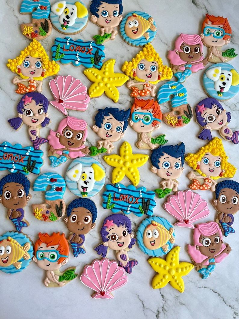 Może przedstawiać: Kolekcja kolorowych, ozdobnych ciasteczek cukrowych z postaciami z dziecięcego programu telewizyjnego Bubble Guppies. Ciasteczka mają kształt ryb, muszli, rozgwiazd i innych obiekt&oacute;w o tematyce oceanicznej. Ciasteczka są ozdobione jasnymi kolorami i skomplikowanymi detalami.