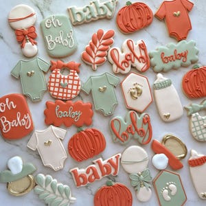 Fall-themed baby shower sugar cookies decorated with royal icing in sage green, burnt orange, ivory, and gold. Designs include pumpkins, “baby” lettering, baby onesies, bottles, bibs, pacifiers, leaves, and gold-accented baby footprints.
