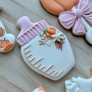 Assorted boho pumpkin baby shower sugar cookies decorated with pumpkins, baby onesie, baby bottle, rainbow, baby feet, “Little Pumpkin,” “Oh Baby,” “Baby Girl,” and “BABY” plaque in soft blush, sage, ivory, and pumpkin orange.