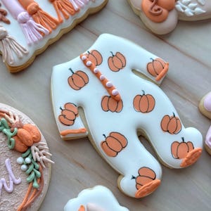 Assorted boho pumpkin baby shower sugar cookies decorated with pumpkins, baby onesie, baby bottle, rainbow, baby feet, “Little Pumpkin,” “Oh Baby,” “Baby Girl,” and “BABY” plaque in soft blush, sage, ivory, and pumpkin orange.