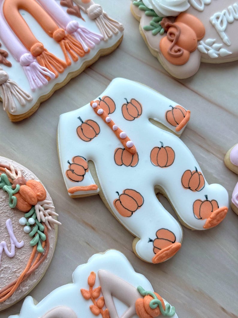 Assorted boho pumpkin baby shower sugar cookies decorated with pumpkins, baby onesie, baby bottle, rainbow, baby feet, “Little Pumpkin,” “Oh Baby,” “Baby Girl,” and “BABY” plaque in soft blush, sage, ivory, and pumpkin orange.