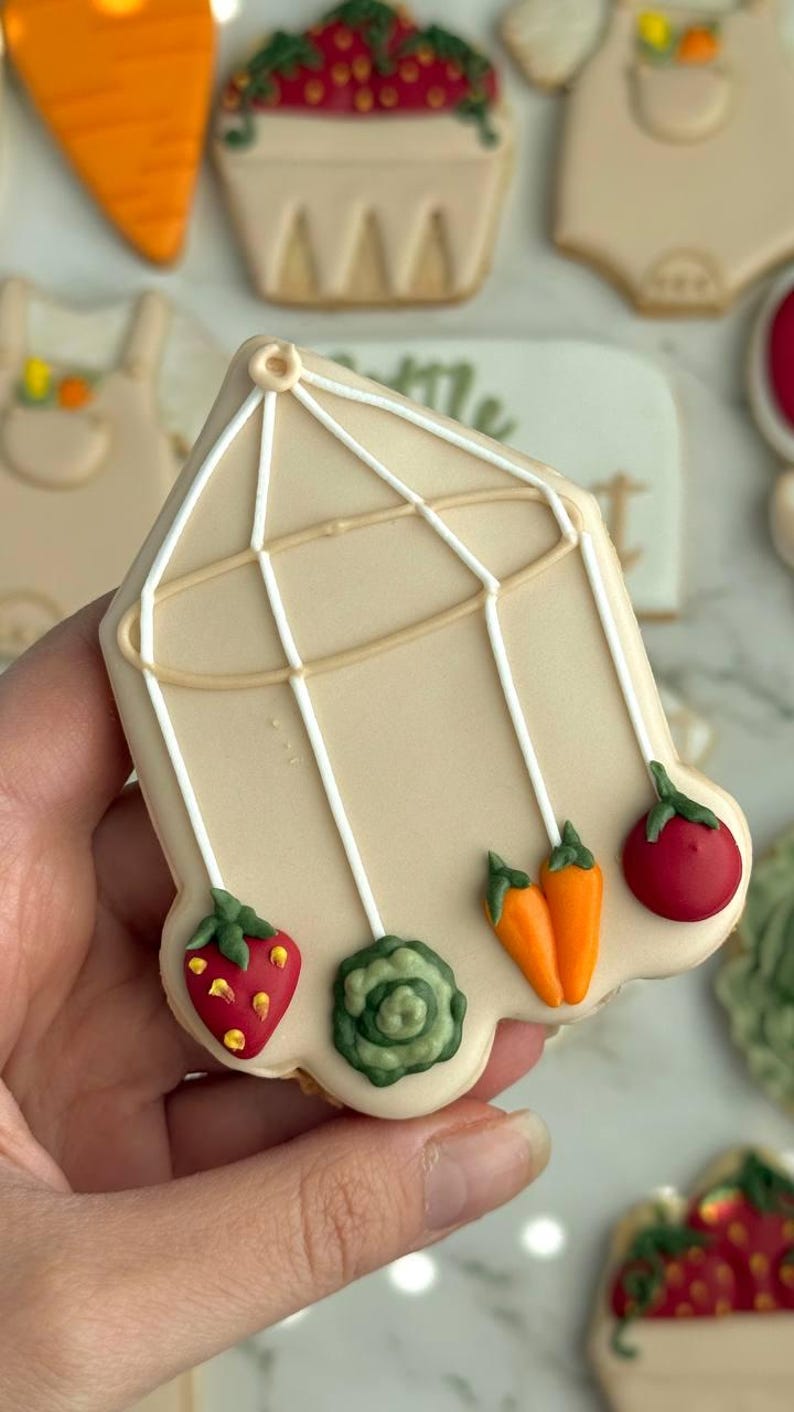 Farmers market-themed baby shower sugar cookies featuring hand-decorated designs like “little sprout,” baby onesies with vegetables, carrots, corn, strawberries, tomatoes, baby bottles, mason jars, leafy greens, and baby mobiles. Adorned with soft green, beige, orange, and red royal icing, these custom cookies are perfect for a “locally grown” or garden baby shower theme, gender-neutral celebrations, and personalized baby gifts.