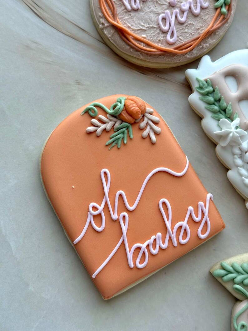 Assorted boho pumpkin baby shower sugar cookies decorated with pumpkins, baby onesie, baby bottle, rainbow, baby feet, “Little Pumpkin,” “Oh Baby,” “Baby Girl,” and “BABY” plaque in soft blush, sage, ivory, and pumpkin orange.