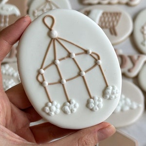 May include: A white cookie decorated with a brown baby mobile with white clouds and beads. The cookie is being held by a hand.