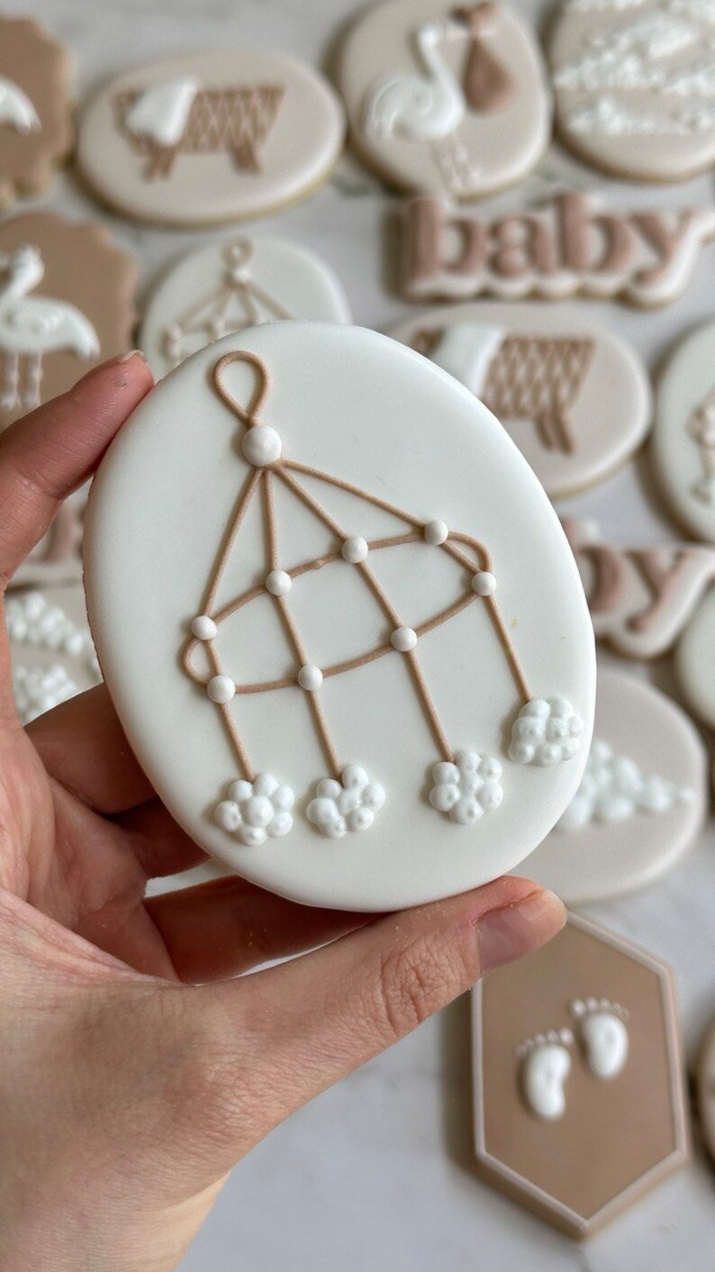 May include: A white cookie decorated with a brown baby mobile with white clouds and beads. The cookie is being held by a hand.
