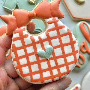 Fall-themed baby shower sugar cookies decorated with royal icing in sage green, burnt orange, ivory, and gold. Designs include pumpkins, “baby” lettering, baby onesies, bottles, bibs, pacifiers, leaves, and gold-accented baby footprints.