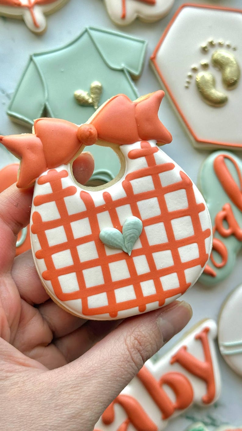 Fall-themed baby shower sugar cookies decorated with royal icing in sage green, burnt orange, ivory, and gold. Designs include pumpkins, “baby” lettering, baby onesies, bottles, bibs, pacifiers, leaves, and gold-accented baby footprints.