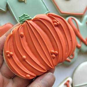 Fall-themed baby shower sugar cookies decorated with royal icing in sage green, burnt orange, ivory, and gold. Designs include pumpkins, “baby” lettering, baby onesies, bottles, bibs, pacifiers, leaves, and gold-accented baby footprints.
