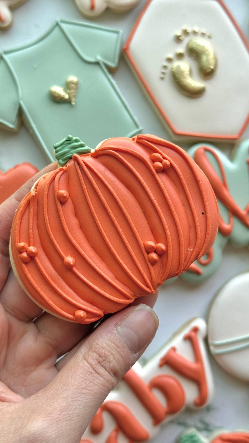 Fall-themed baby shower sugar cookies decorated with royal icing in sage green, burnt orange, ivory, and gold. Designs include pumpkins, “baby” lettering, baby onesies, bottles, bibs, pacifiers, leaves, and gold-accented baby footprints.
