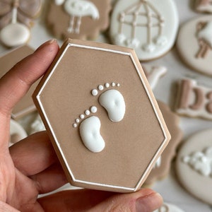 May include: A hexagonal brown sugar cookie with white icing depicting two baby footprints. The cookie is decorated with a white outline.