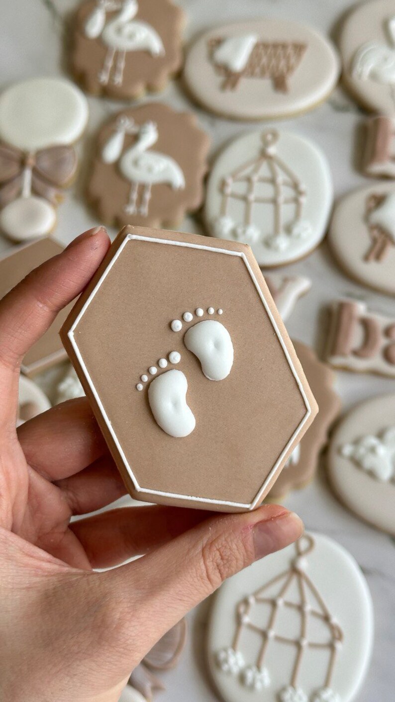 May include: A hexagonal brown sugar cookie with white icing depicting two baby footprints. The cookie is decorated with a white outline.