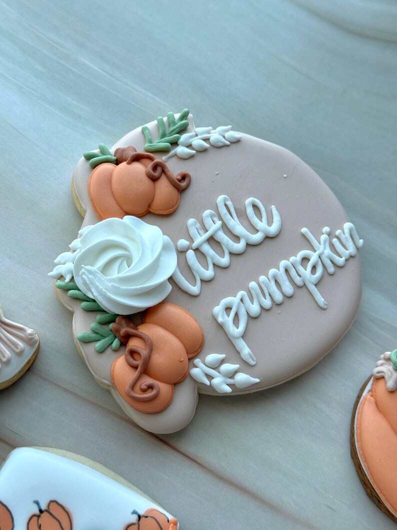 Assorted boho pumpkin baby shower sugar cookies decorated with pumpkins, baby onesie, baby bottle, rainbow, baby feet, “Little Pumpkin,” “Oh Baby,” “Baby Girl,” and “BABY” plaque in soft blush, sage, ivory, and pumpkin orange.