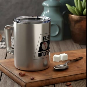May include: A brushed stainless steel mug with a clear lid and a handle, featuring a black graphic design with the text ALPHA MOONBASE and illustrations of the Earth and the moon. The mug is cylindrical with a flat base and a smooth metallic finish.